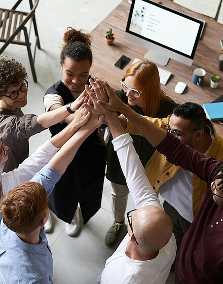 Lachende Kolleginnen und Kollegen strecken beim Teambuilding-Event im Kreis die Hände gemeinsam in die Mitte