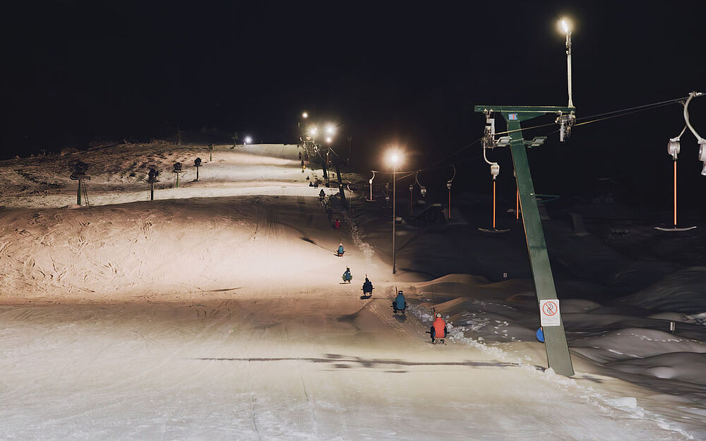 eleuchtete Skipiste bei Nacht mit Schlepplift, der rodelnde Personen bergauf zieht