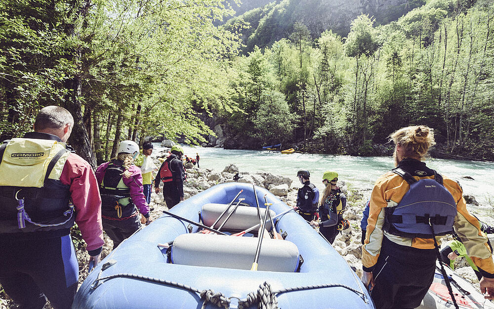 Teilnehmende tragen ein Raftingboot zum Fluss, im Hintergrund fließt die Isar