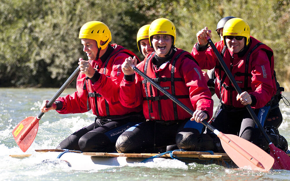 Personen mit Neoprenanzügen, Schwimmwesten und Helmen paddeln auf selbst gebautem Floß die Isar hinunter
